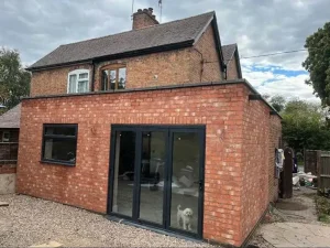 Newly completed rear extension with matching red brickwork and dark grey aluminium bi-fold doors at a home in Brandon, Coventry, with a small dog visible inside.